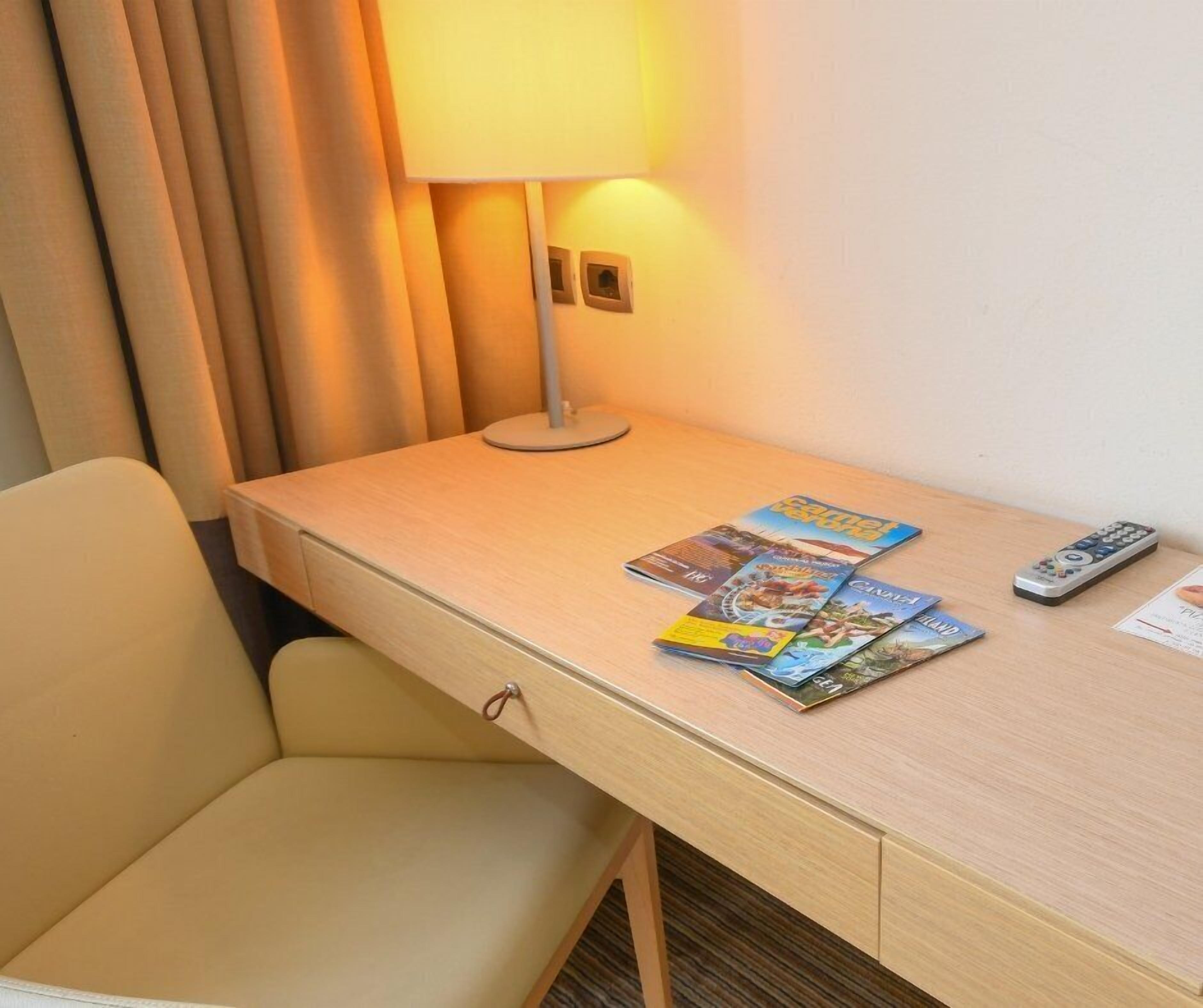 Une chambre d’hôtel avec un bureau en bois, une lampe, une télécommande et un magazine.
