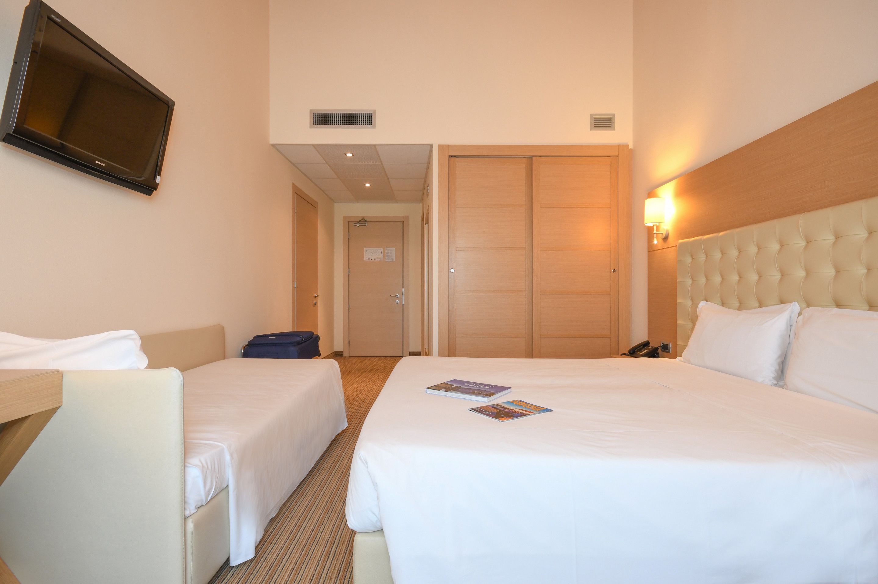 A hotel room with two beds, a TV mounted on the wall, a bedside lamp, and a door leading to another room.