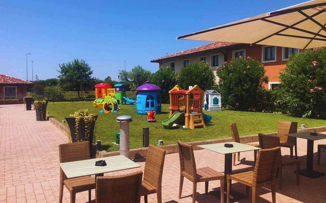 Un área exterior con asientos de mimbre y mesas, un espacio de juegos infantiles con estructuras coloridas y un edificio con tejado de tejas rojas al fondo.