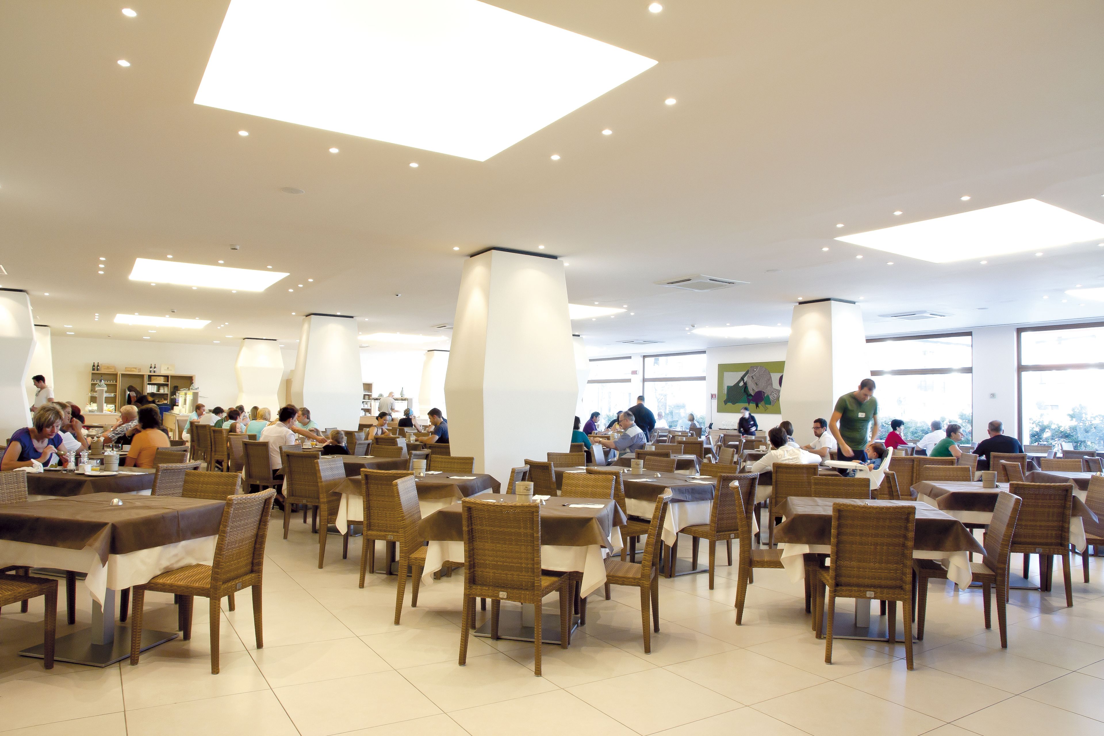 A spacious dining area with multiple tables and chairs, large windows, and a high ceiling with recessed lighting.