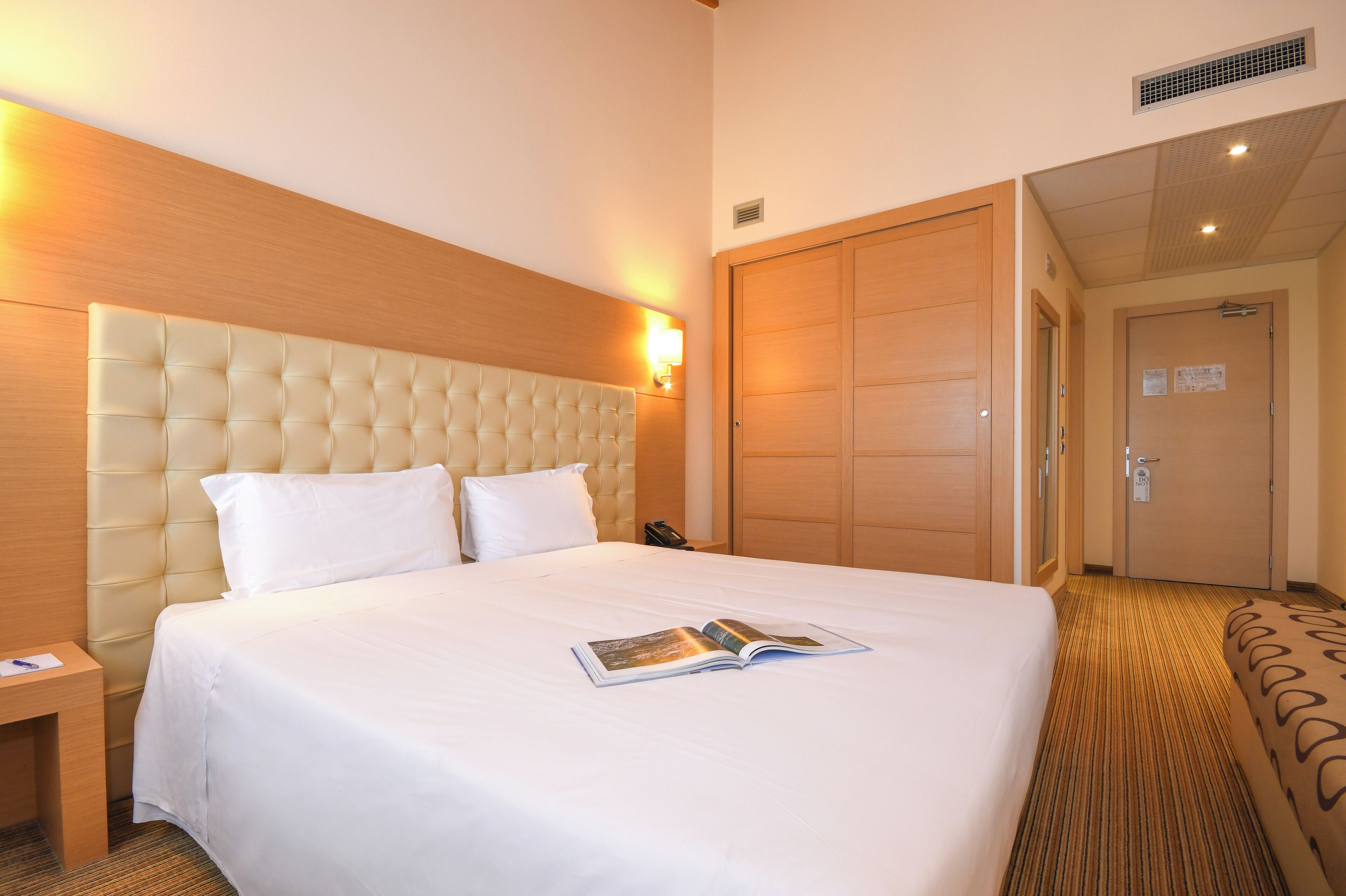 A hotel room with a large bed, a wooden headboard, a bedside table, and a door leading to another room.