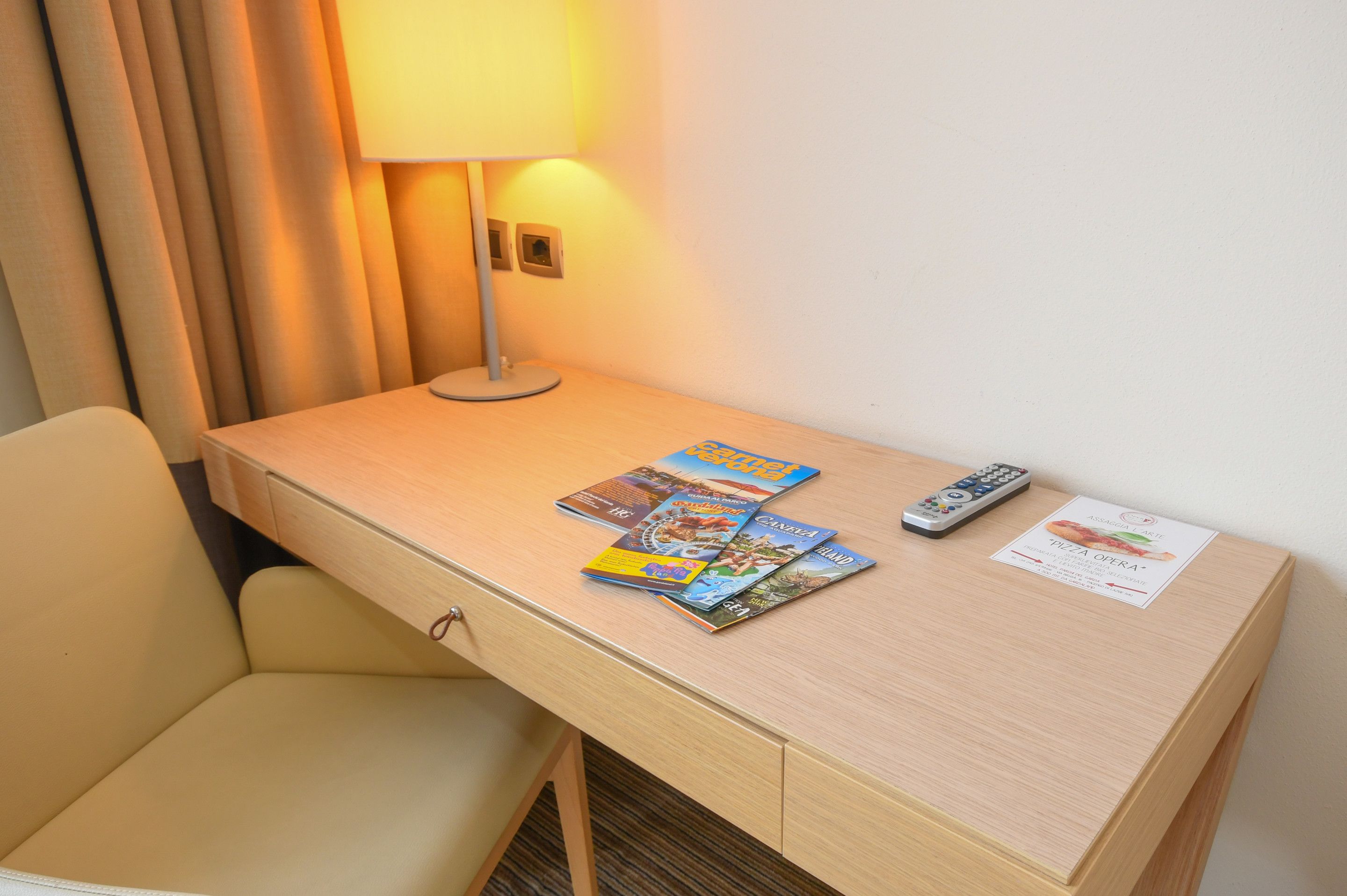 A hotel room with a wooden desk, a lamp, a remote control, and a menu card.