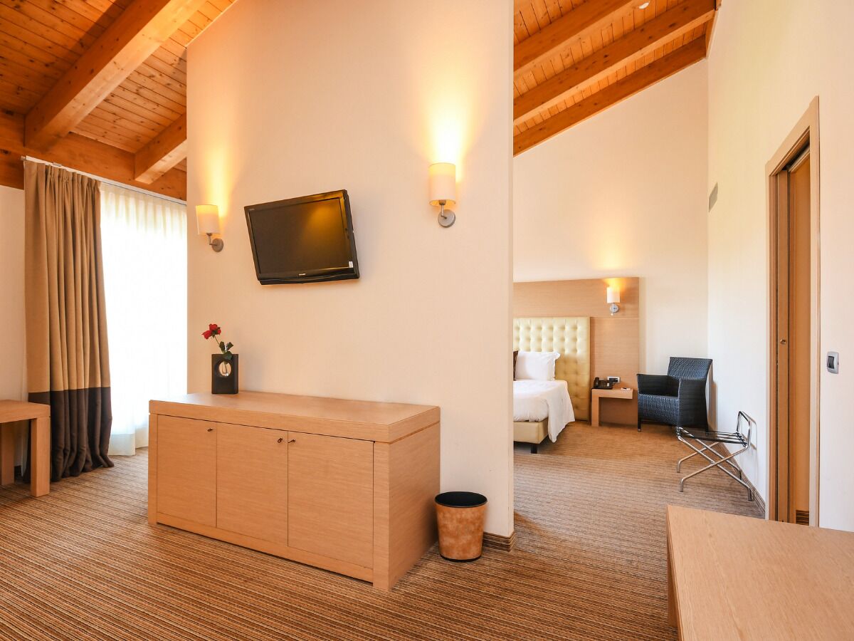 A hotel room with a wooden ceiling, a flat-screen TV mounted on the wall, a bed with a white headboard, a bedside table, a chair, and a small desk.