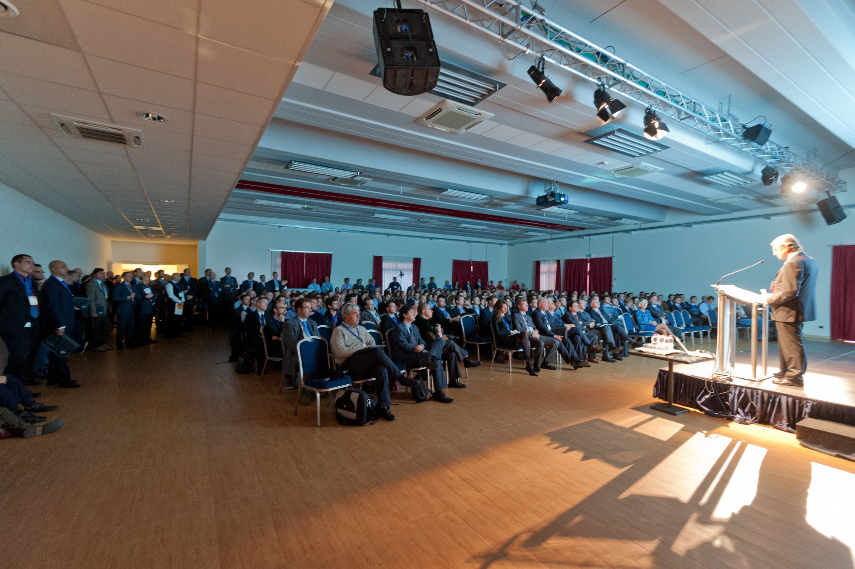 Een ruime zaal met rijen stoelen gericht op een podium waar een spreker een publiek toespreekt.