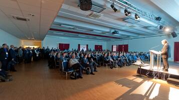 Een ruime zaal met rijen stoelen gericht op een podium waar een spreker een publiek toespreekt.