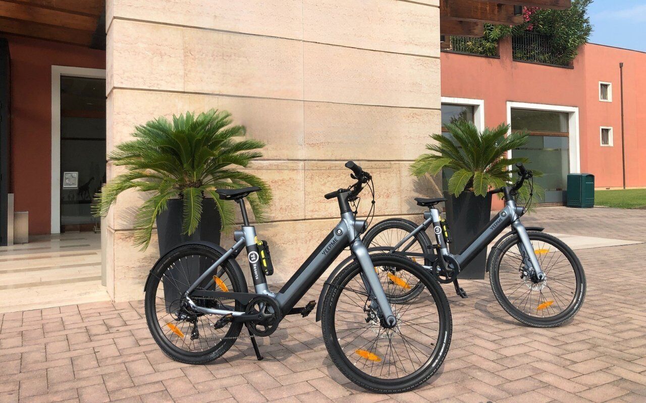 Two bicycles parked against a building with a tiled facade and potted plants.