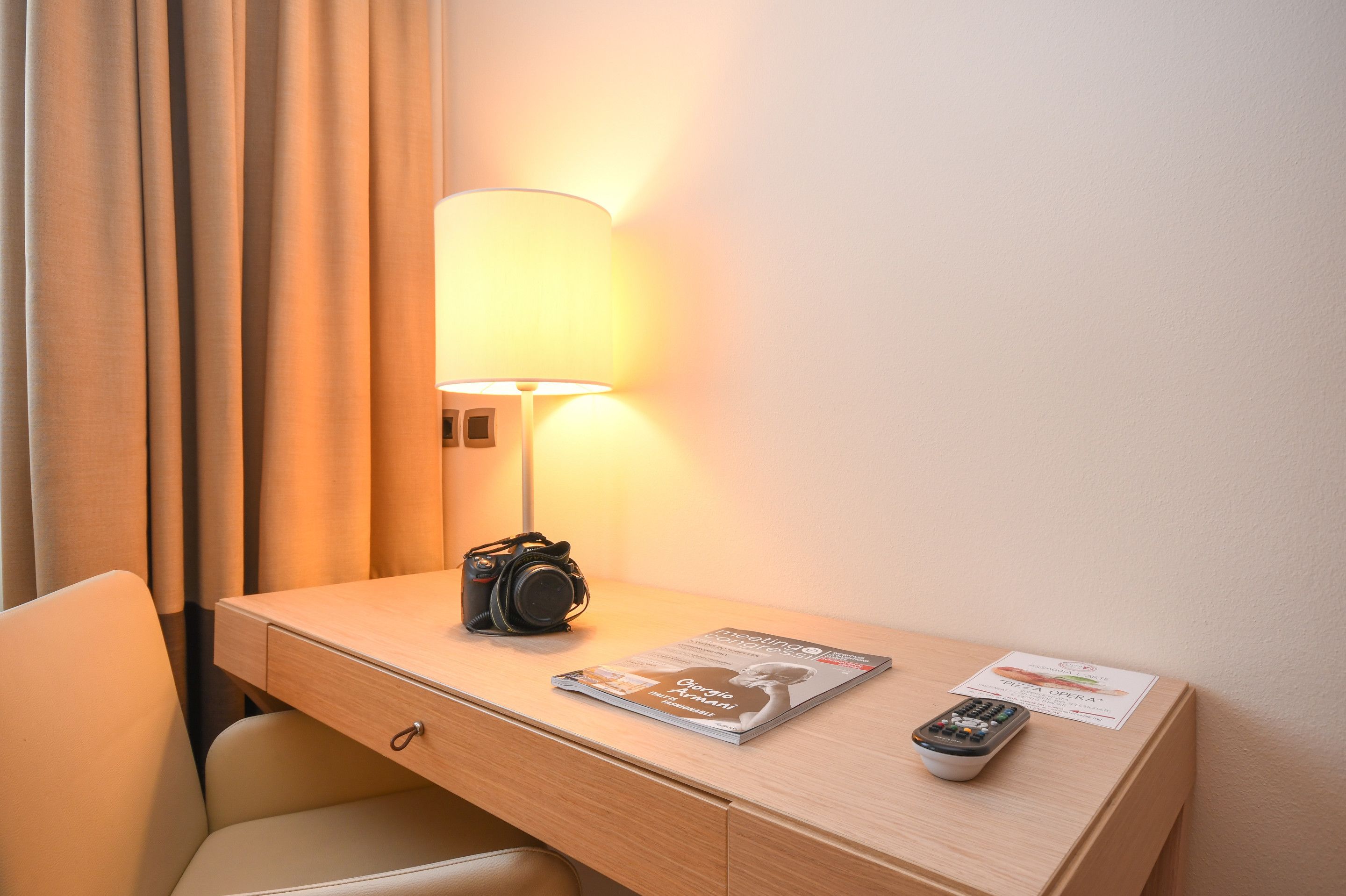Habitación de hotel con un escritorio de madera, una lámpara, una cámara, un control remoto y una revista.