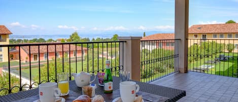 Balcón con artículos de desayuno, como cruasanes, café y jugo, con vistas a un paisaje de edificios y follaje.