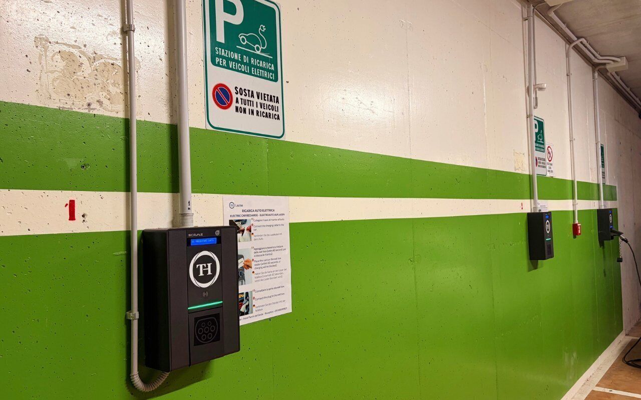 Un área de estacionamiento con estaciones de carga para vehículos eléctricos, un letrero que indica una zona de estacionamiento y un dispositivo montado en la pared con una etiqueta.