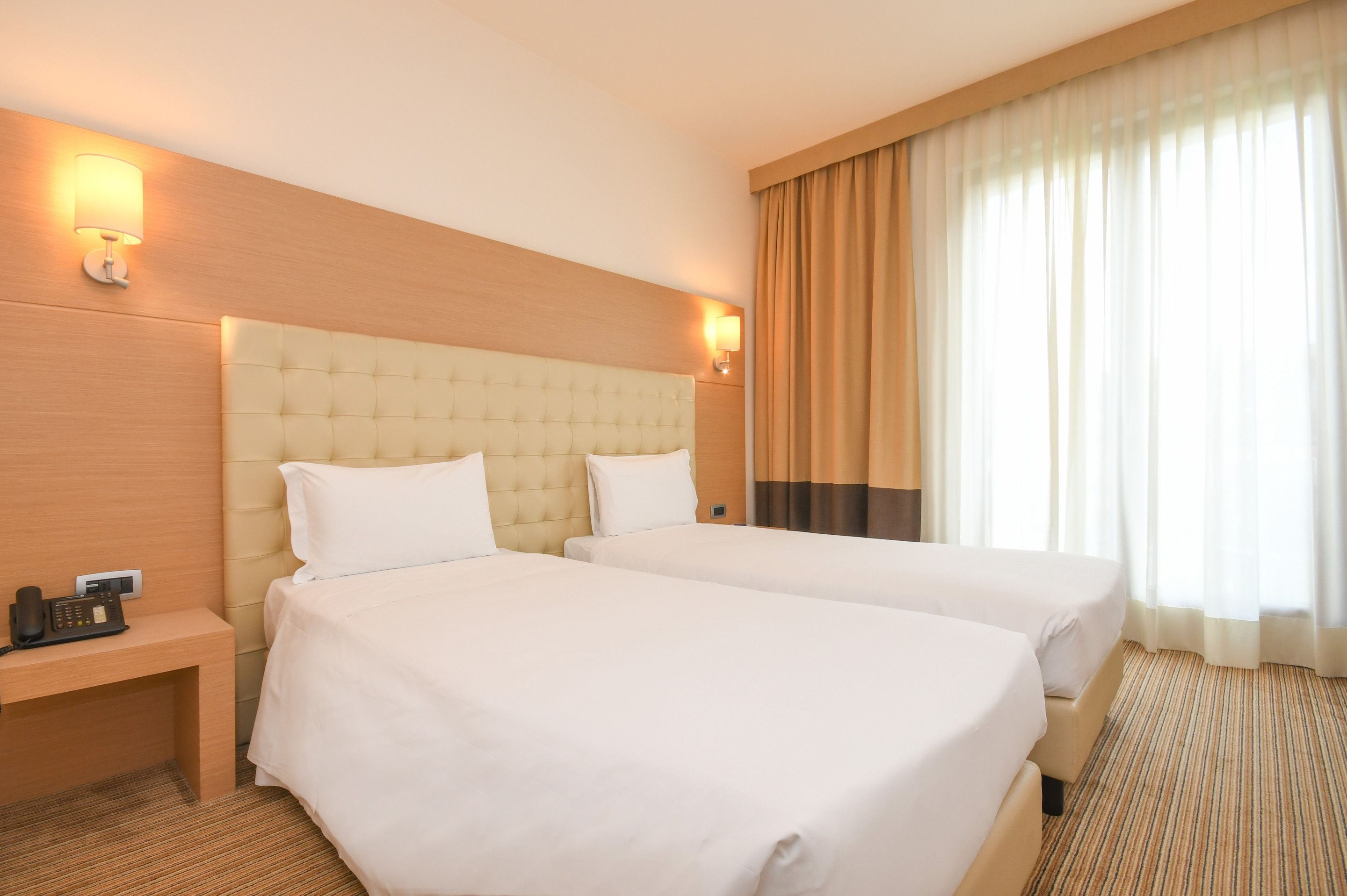 A hotel room with two beds, a wooden headboard, bedside lamps, a telephone, and a window with curtains.