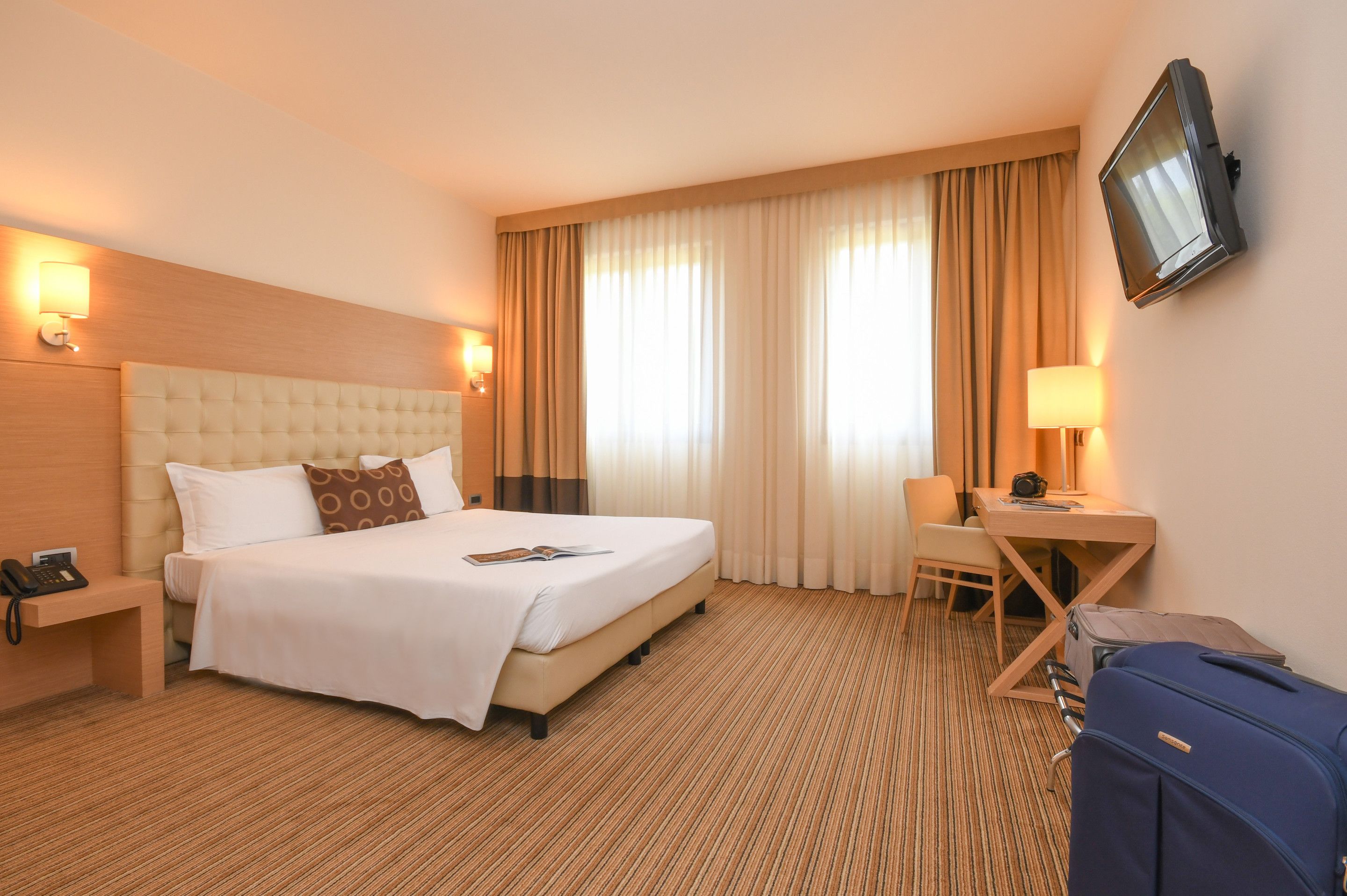 A hotel room with a large bed, a desk, a chair, a TV, and a suitcase.