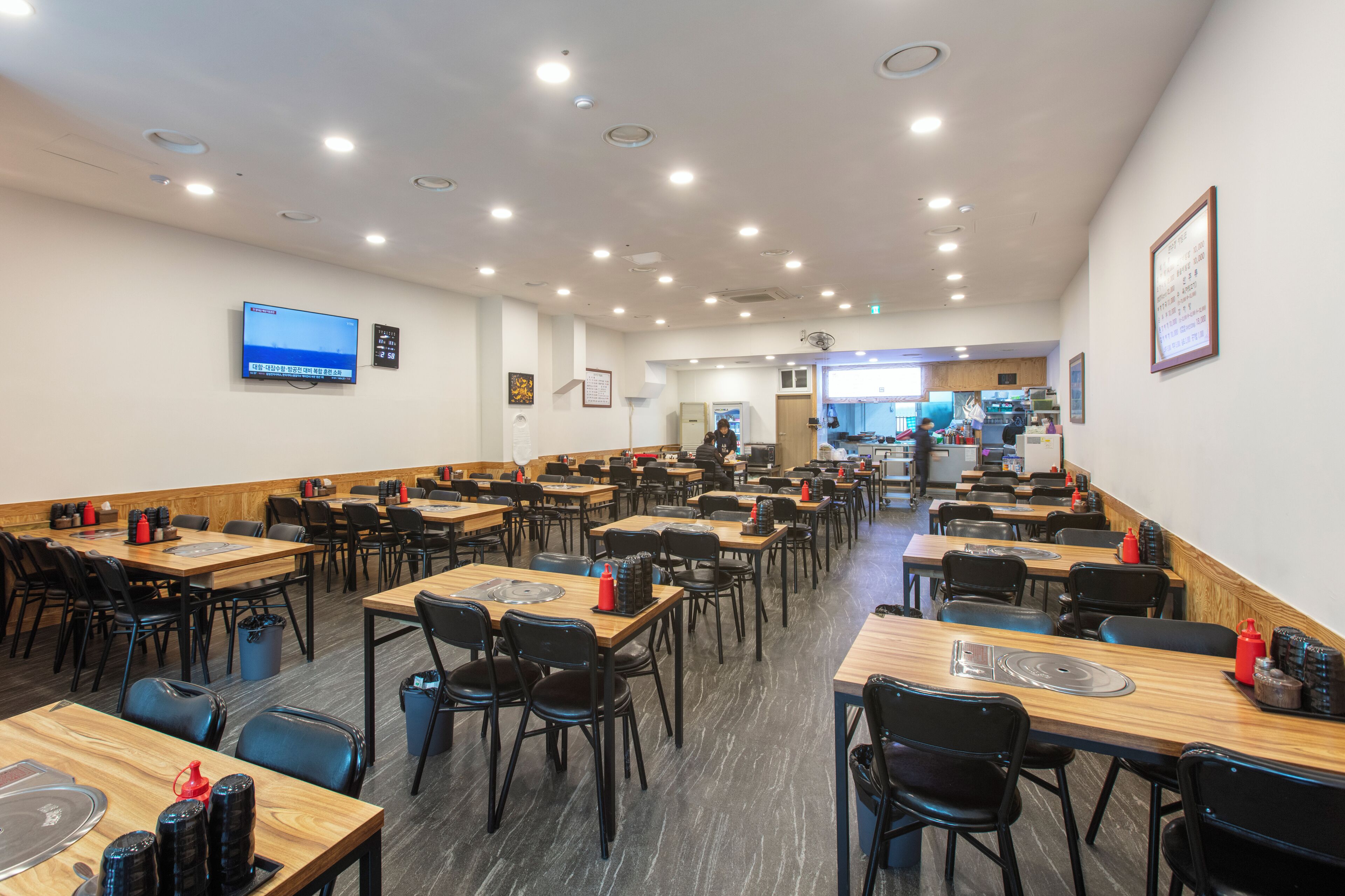 A spacious dining area with multiple tables and chairs, a television mounted on the wall, and a kitchen area in the background.