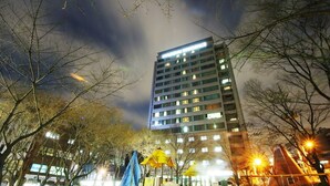 Un hôtel moderne à plusieurs étages, avec une enseigne bien visible au sommet, et un hall d’entrée bien éclairé agrémenté d’arbres et de sièges extérieurs.