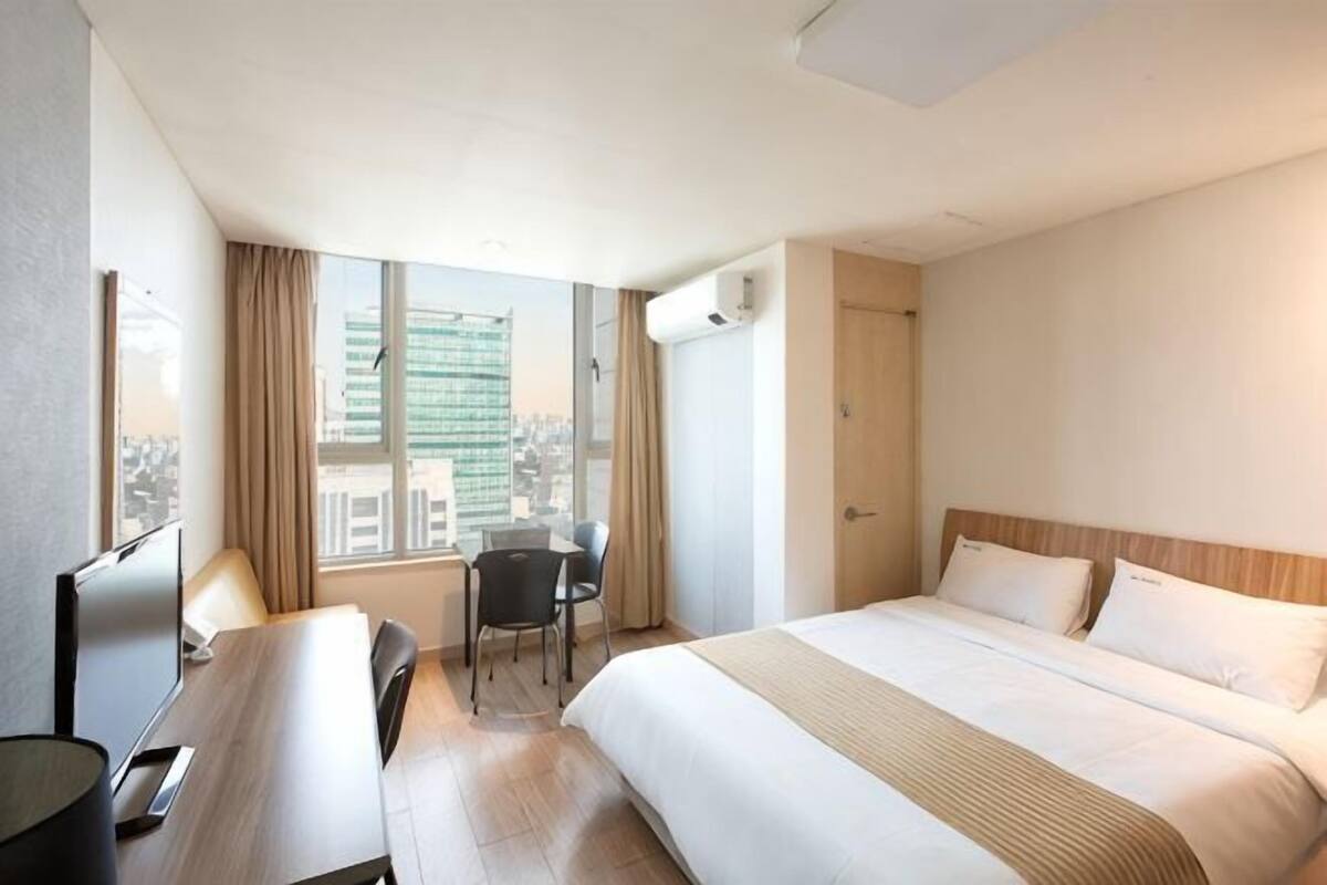 Ein Hotelzimmer mit Bett, Schreibtisch, Sessel, Fernseher und Fenster mit Blick auf die Stadt.