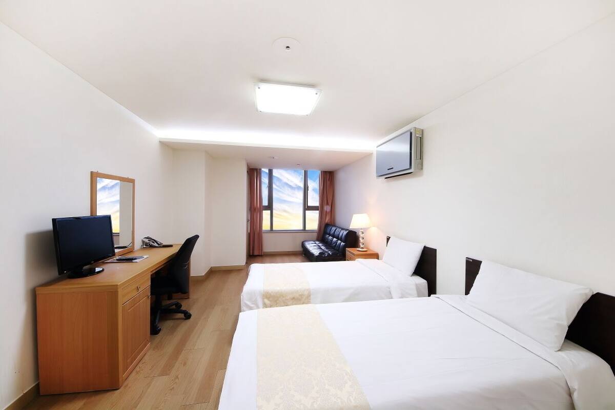 Ein Hotelzimmer mit zwei Betten, einem Schreibtisch mit Computer, einem Fernseher und einem Fenster mit Vorhängen.