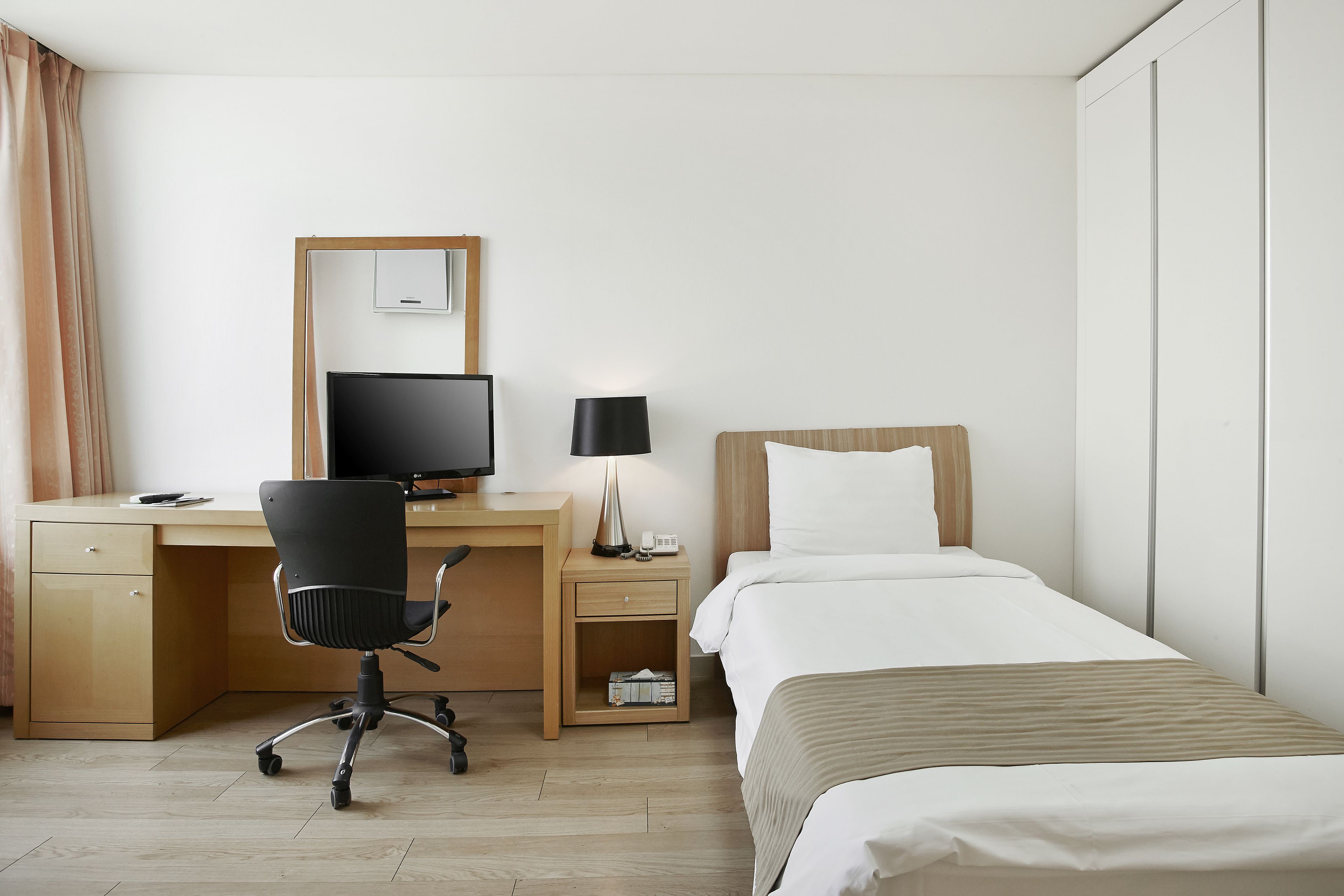 Quarto de hotel com uma cama, uma escrivaninha com computador, uma cadeira, um abajur e um guarda-roupa.
