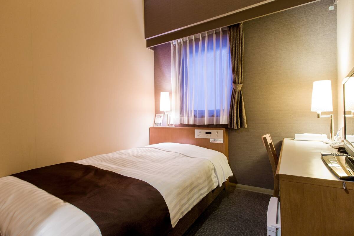 A hotel room with a bed, a desk with a computer, a chair, and a window with curtains.