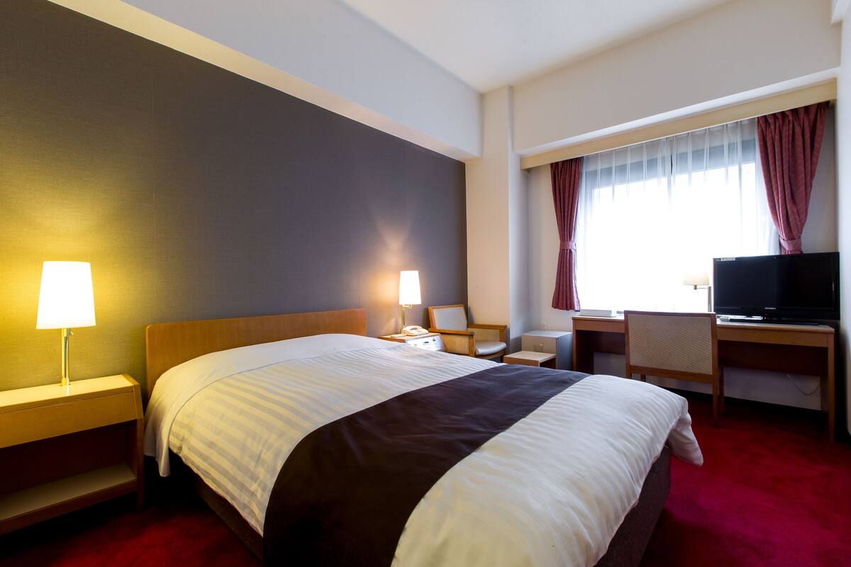 A hotel room with a large bed, bedside lamps, a desk with a chair, a television, and a window with curtains.