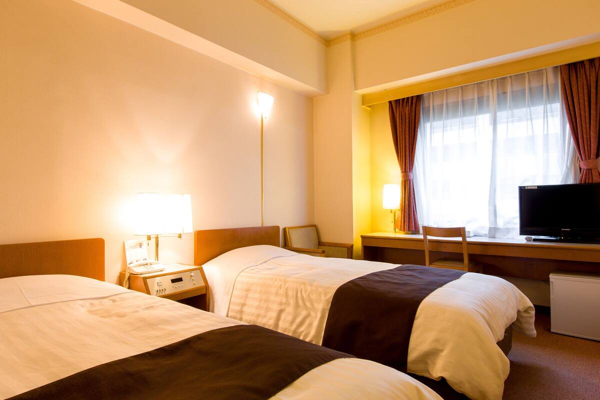 A hotel room with two beds, a desk with a television, and a window with curtains.