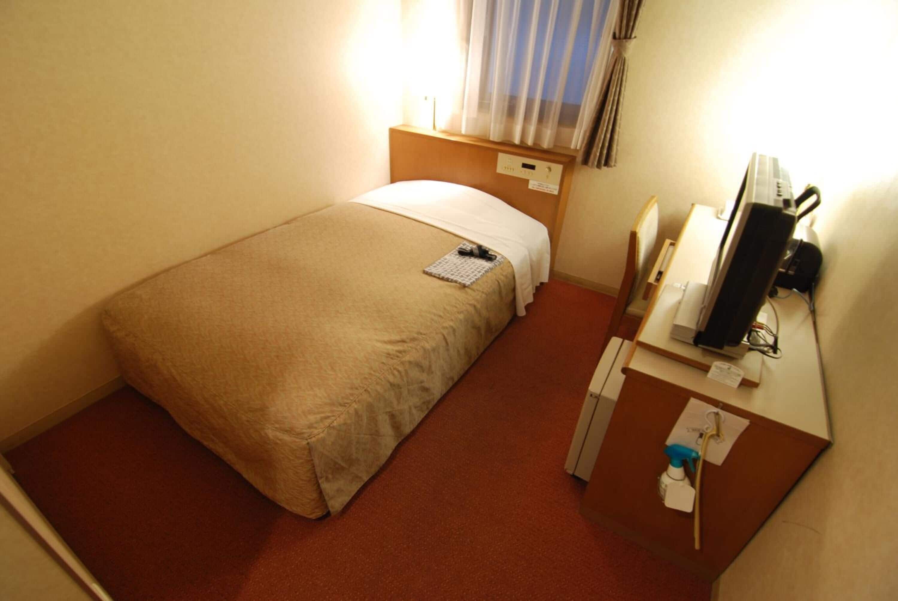 A hotel room with a single bed, a desk with a computer, and a small table with a spray bottle.