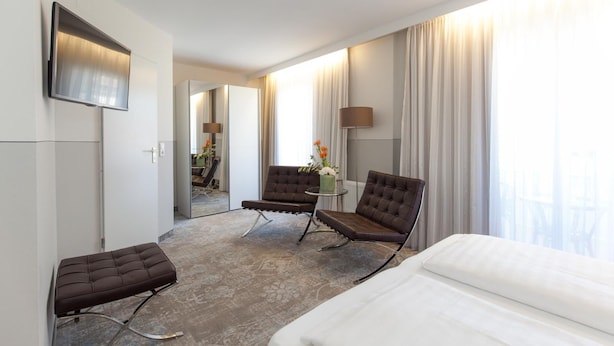 Ein Hotelzimmer mit Bett, zwei Sesseln, einem kleinen Tisch mit Blumenvase, einem Flachbildfernseher und einem großen Fenster mit Vorhängen.