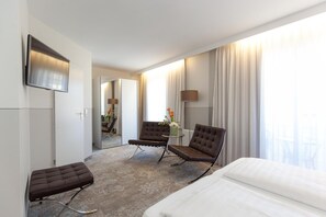 Une chambre d’hôtel comprenant un lit, deux fauteuils, une petite table avec un vase de fleurs, un téléviseur à écran plat et une grande fenêtre avec des rideaux.