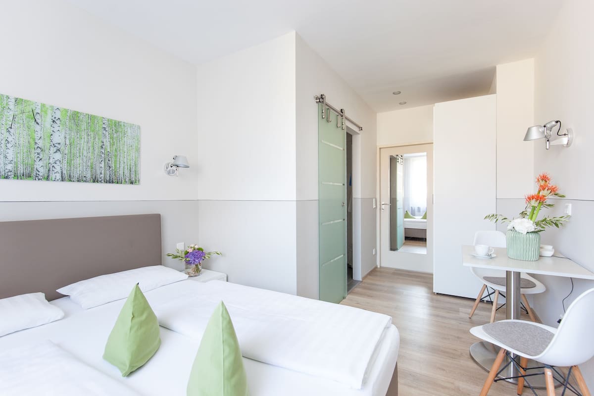 Une chambre d’hôtel moderne, équipée d’un lit, d’un bureau et d’une chaise. On y trouve une porte donnant sur une autre pièce et une lampe fixée au mur.