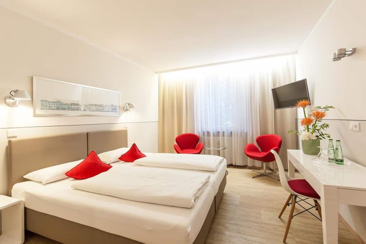 Une chambre d’hôtel avec un lit, un bureau, une chaise, une télévision et une fenêtre avec des rideaux.