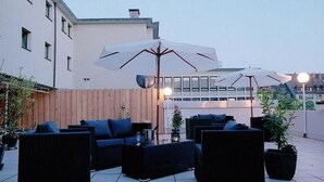 Ein Außenbereich mit schwarzer Rattanmöbel und weißen Sonnenschirmen, umgeben von einem Holzzaun und Pflanzkübeln.