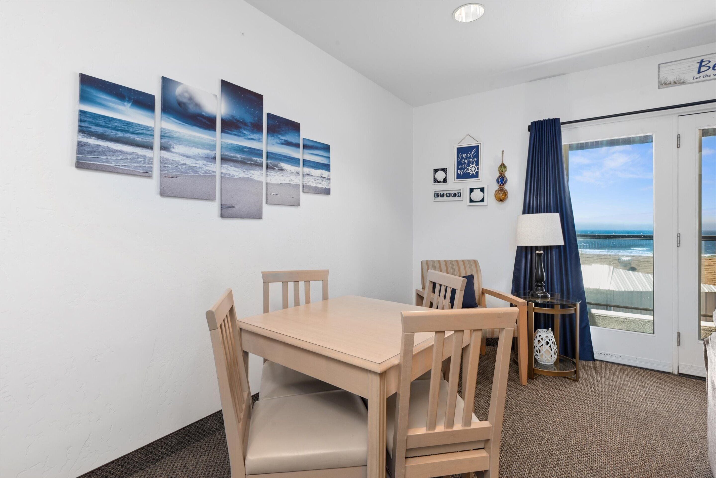A room with a wooden table and chairs, a small side table with a lamp, a wall with framed artwork, and a door leading to a balcony with a view of the ocean.