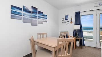 A room with a wooden table and chairs, a small side table with a lamp, a wall with framed artwork, and a door leading to a balcony with a view of the ocean.