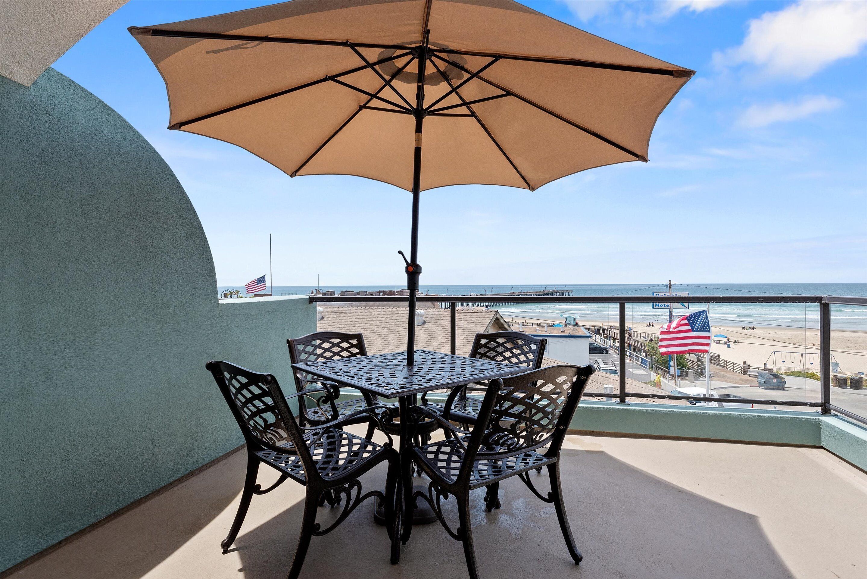A balcony with a table and chairs, an umbrella, and a view of the beach and ocean.