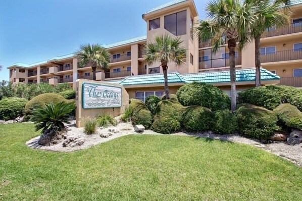 A hotel with a sign reading "The Oasis." It features a multi-story building with balconies and a well-maintained garden with palm trees and shrubs.