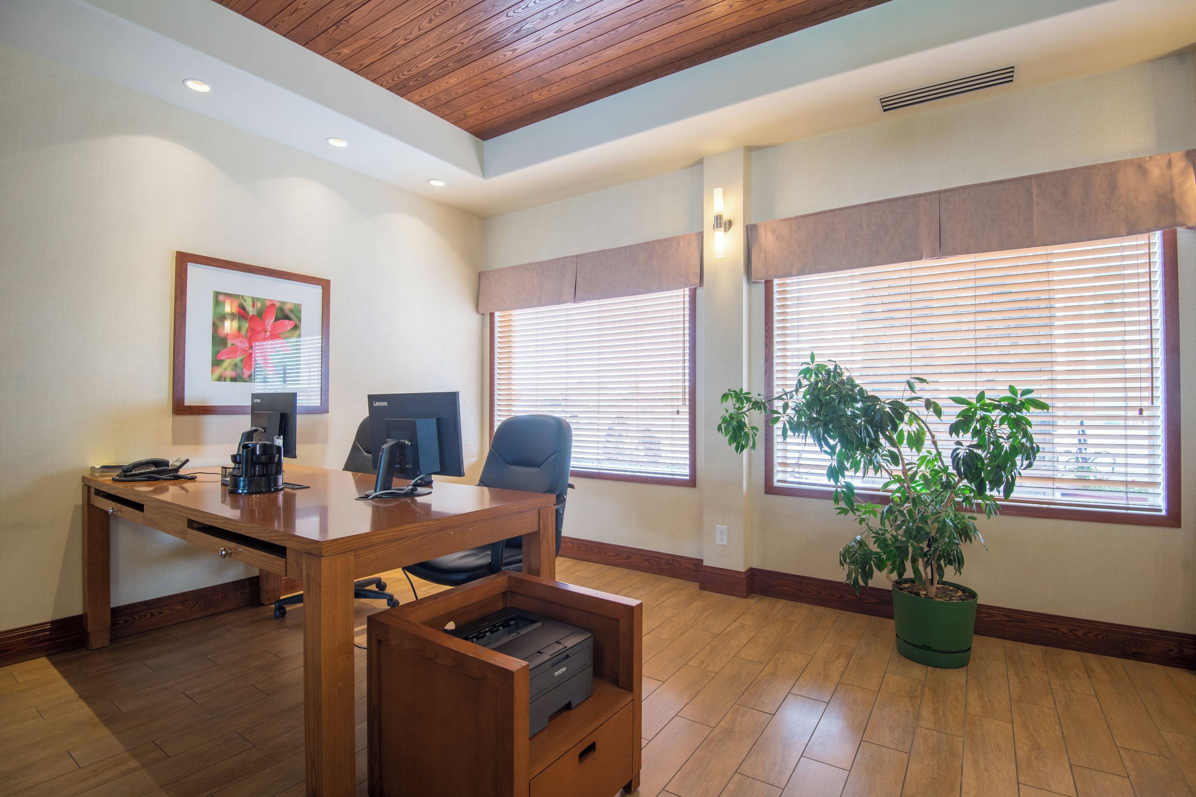 A modern office space with a wooden desk, two computer monitors, a printer, a telephone, and a framed picture on the wall.