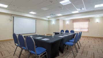 Un salón de conferencias con una larga mesa, sillas azules, una pantalla de proyector y una ventana con persianas.