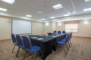 Una sala de conferències amb una taula llarga, cadires blaves, una pantalla de projector i una finestra amb persianes.