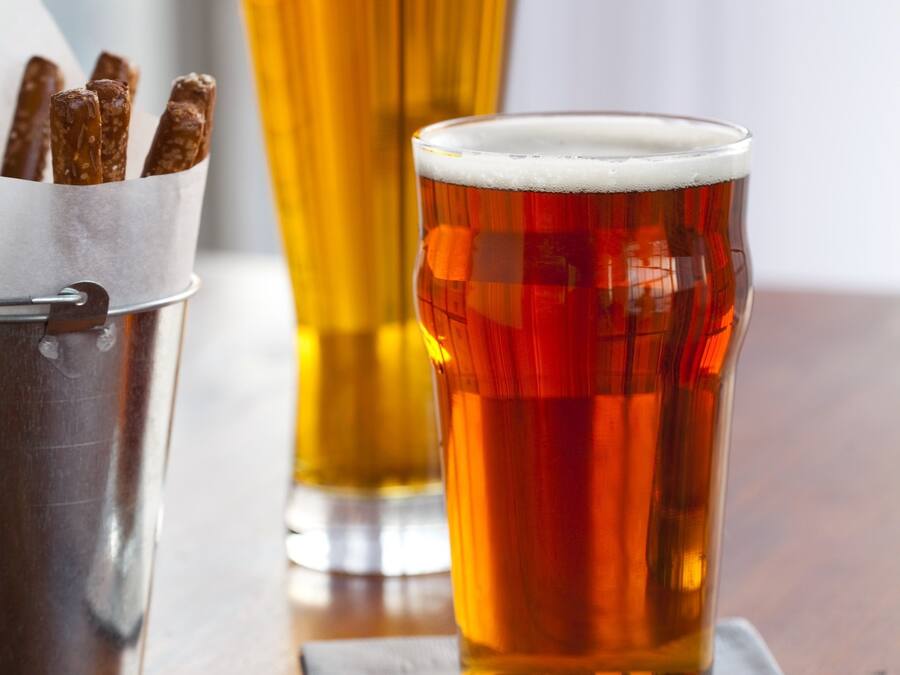 Two glasses of beer on a wooden table with a metal container of pretzels.