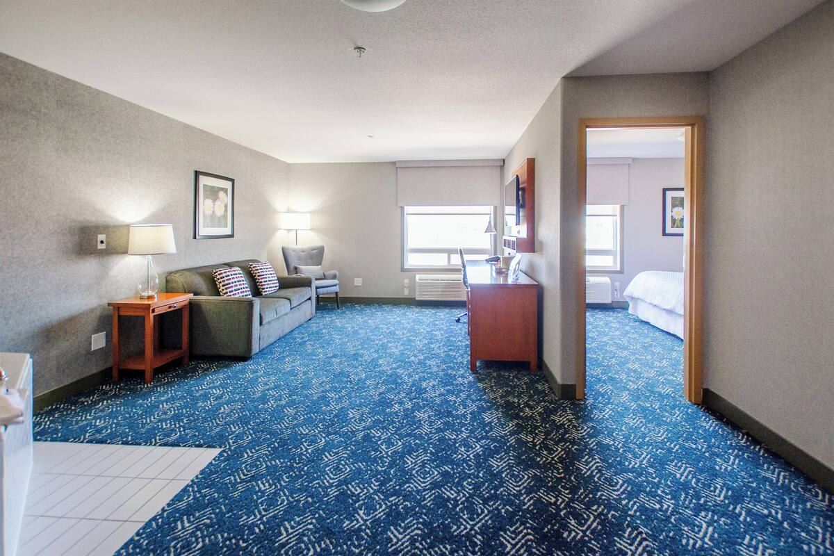 A hotel room with a blue patterned carpet, a sofa, a small table, a lamp, and a bed in the background.