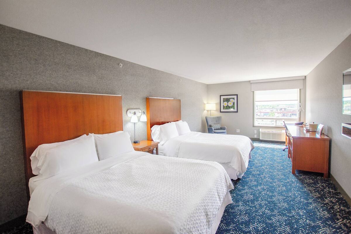 A hotel room with two beds, a desk, a chair, a lamp, and a window with a view.