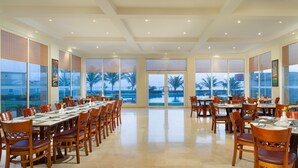A spacious dining area with multiple tables and chairs, large windows with views of palm trees and a pool, and a modern ceiling design. - Masira Island Resort (Masirah)