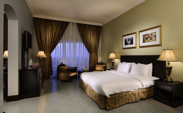 A hotel room with a large bed, two bedside tables with lamps, a small table with a vase, a chair, a TV, and a window with curtains.