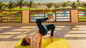 A person performing a yoga pose on a yellow mat with a view of a beach and palm trees in the background. - Masira Island Resort (Masirah)