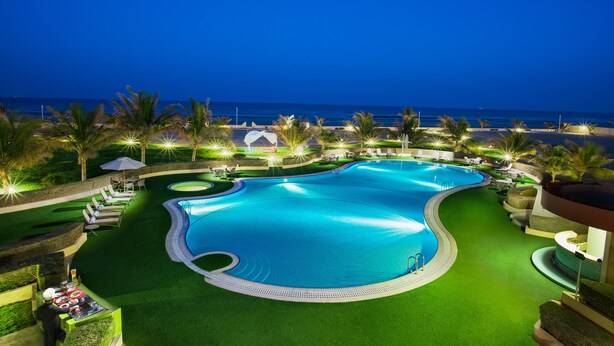 A large, curved swimming pool with a surrounding deck, palm trees, and lounge chairs. - Masira Island Resort (Masirah)