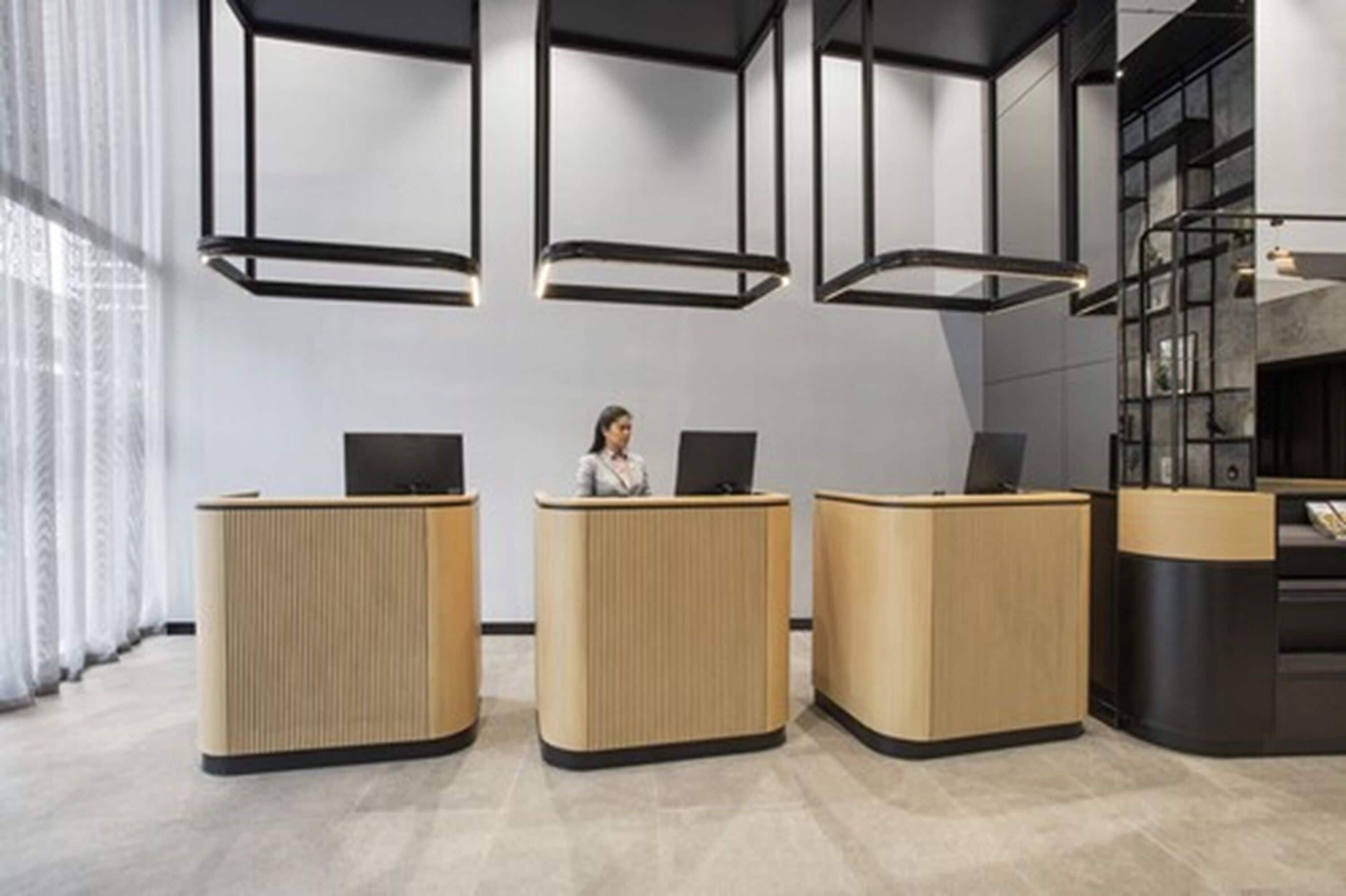 Un espace d’accueil comprenant trois comptoirs de réception en bois, une personne assise au comptoir central et un luminaire moderne au-dessus.