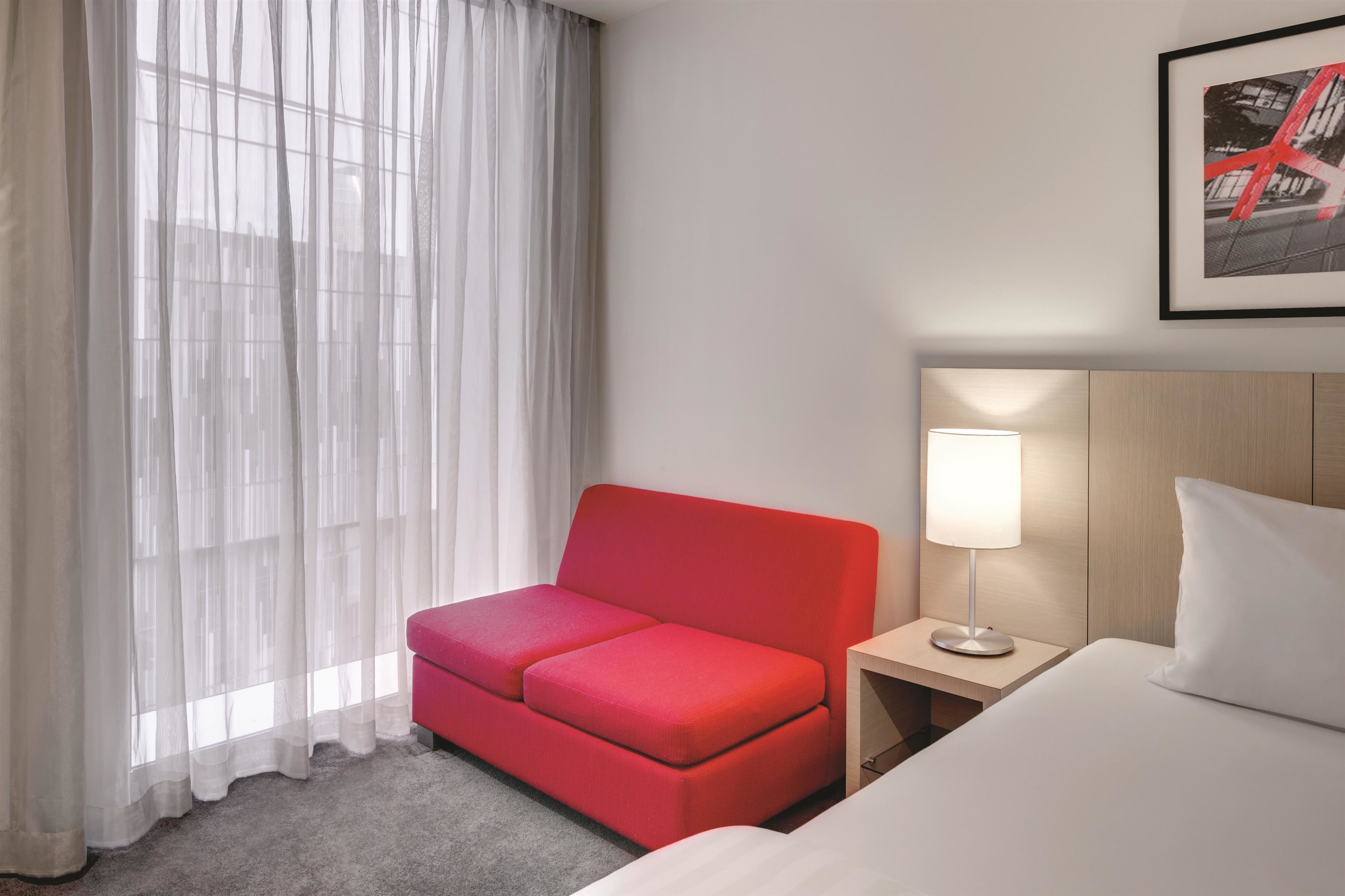 Una habitación de hotel con un sofá rojo, una cama blanca, una mesita de noche con una lámpara y un cuadro en la pared.