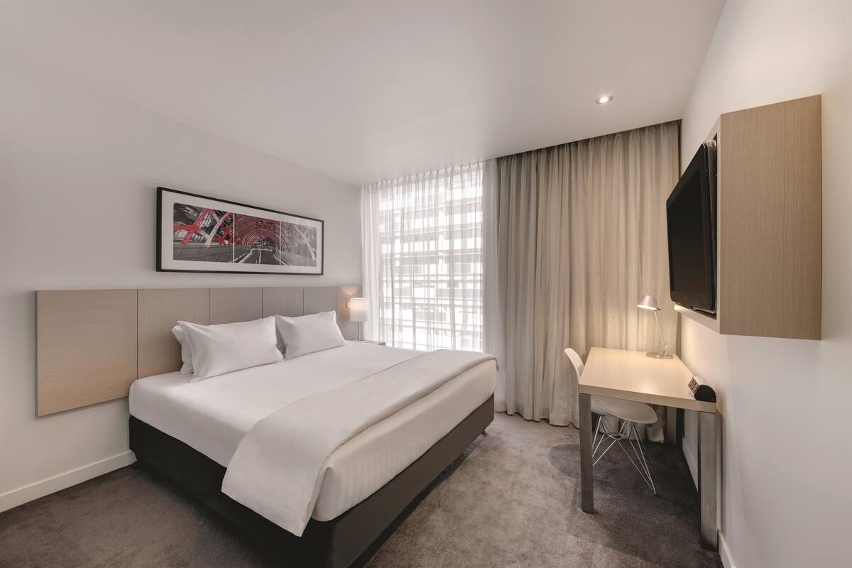 Ett hotellrum med en säng, ett skrivbord och ett stort fönster med gardiner.