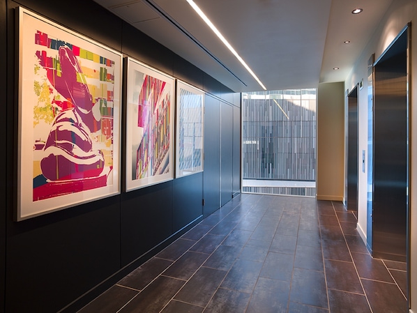 A hallway with three framed artworks on the wall, a modern elevator, and a glass door leading to a bright area.