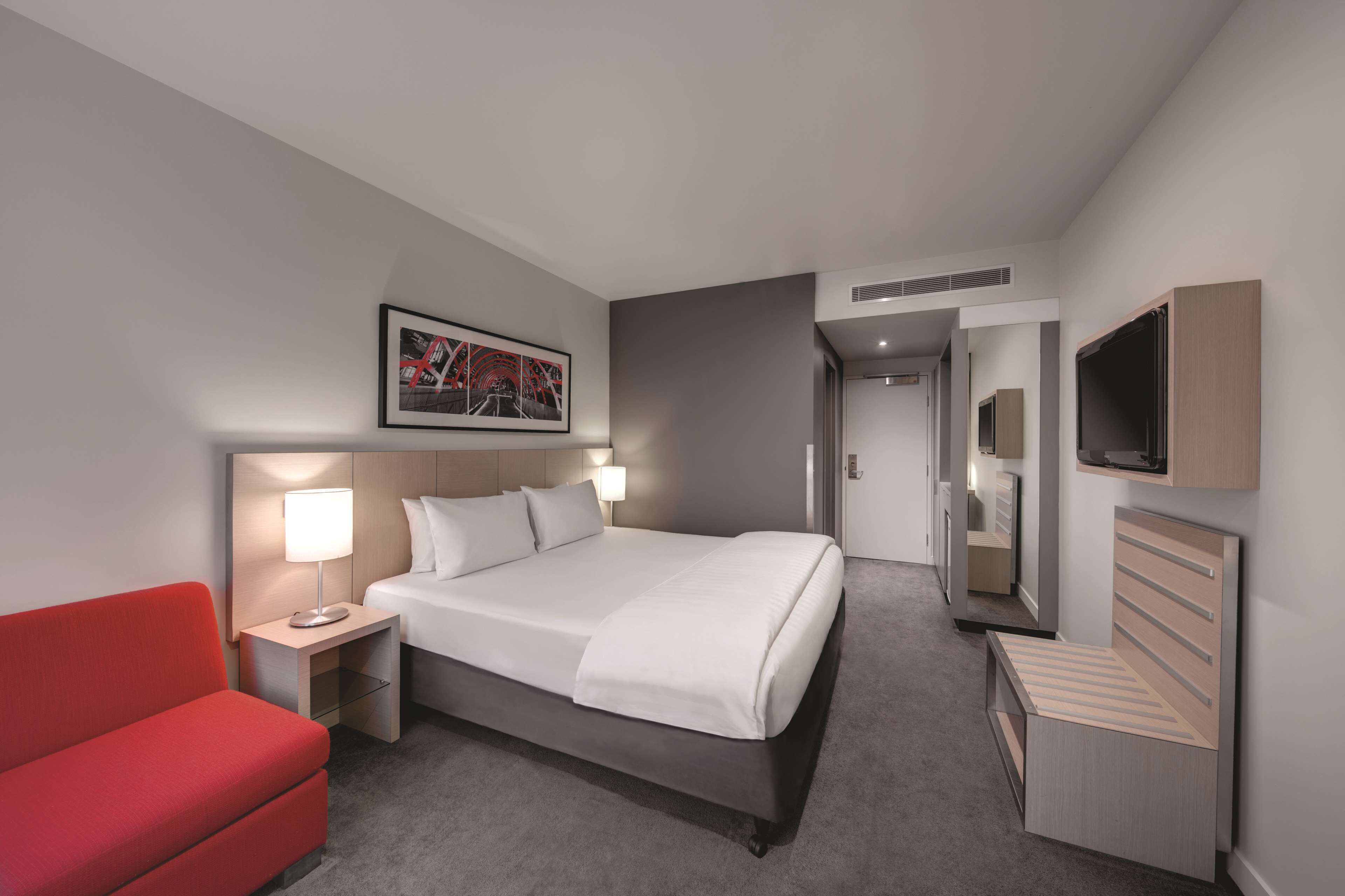 Habitación de hotel con una cama, una silla roja, una mesita de noche con lámpara, un televisor montado en la pared y un cuadro enmarcado sobre la cama.