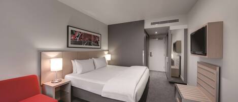 Habitación de hotel con una cama, una silla roja, una mesita de noche con lámpara, un televisor montado en la pared y un cuadro enmarcado sobre la cama.