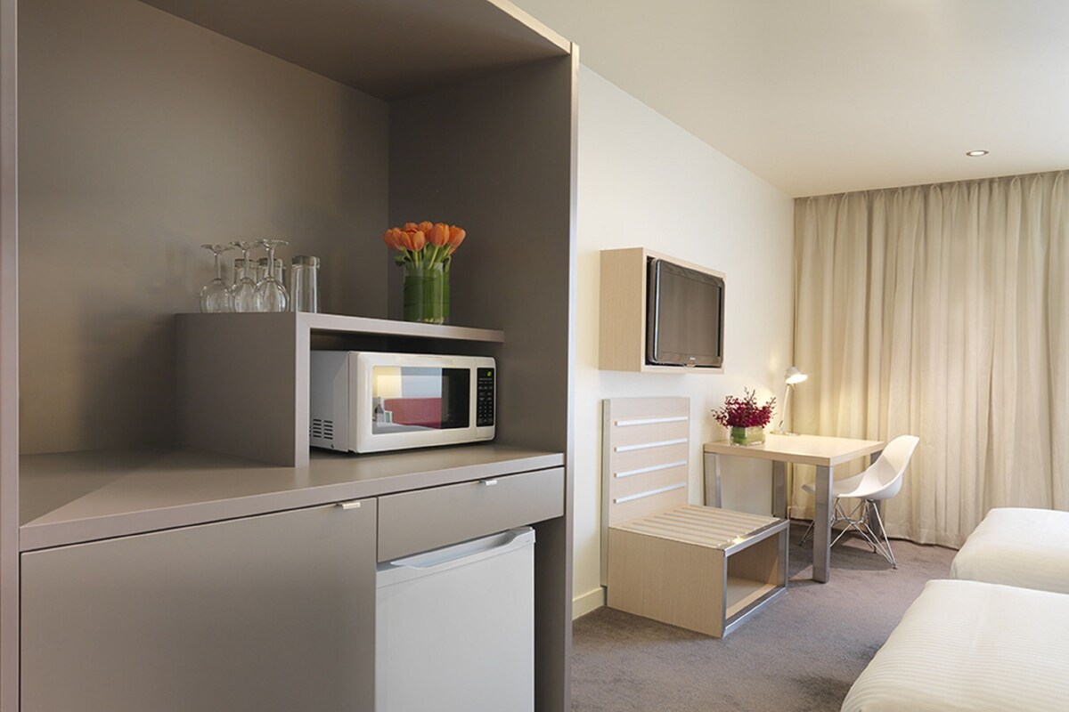 Hotelkamer met kitchenette, bed, bureau met stoel, televisie en raam met gordijnen.
